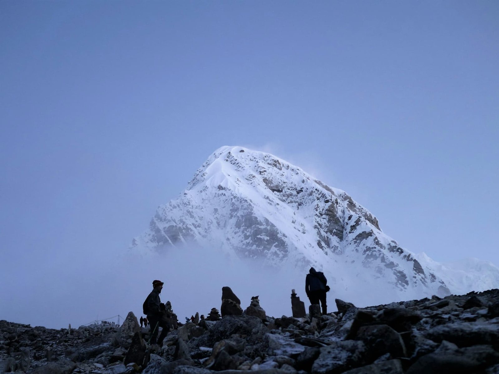 Marcheurs en silhouette parmi les cairns du camp de base de l'Everest, à l'aube, face à un sommet enneigé.
